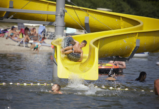 Niño en un flotador baja por tobogán amarillo al lago en TerSpegelt WaterLodges Brabant, camping glamping.