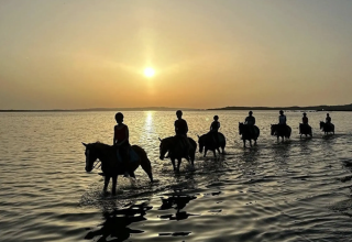 Sunset at Lavita Camp Cagliari, Sardinia, people riding horses through shallow water near glamping tents.