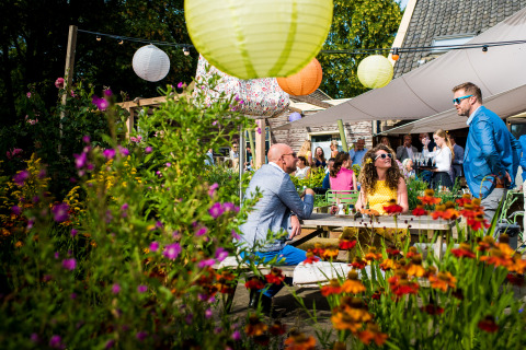 Buitenfeest bij De Vreemde Vogel glamping met kleurrijke lampionnen, bloemen en lachende gasten in de zon.