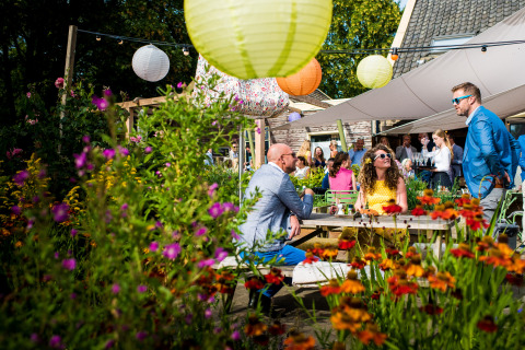 Udendørs glamping-fest på De Vreemde Vogel med farverige lanterner, blomster og glade gæster i solen.