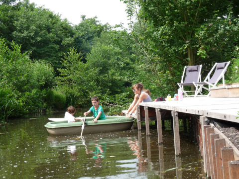 Kinder rudern in einem kleinen Boot neben einem Steg, mit Erwachsenen, Stühlen und Natur in Holland.