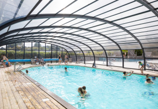 Indoor pool with transparent roof and people swimming and relaxing at Huttopia Les Falaises - Glamping Normandië.