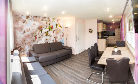 Moderna sala de glamping con mural floral, sofás grises, mesa de comedor y cocina abierta en Camping Teichmann.