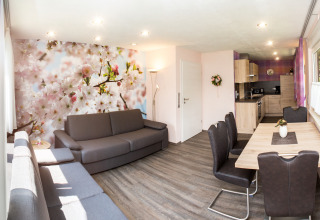 Moderne glamping-stue med blomsterfototapet, grå sofaer, spisebord og åbent køkken ved Camping Teichmann.