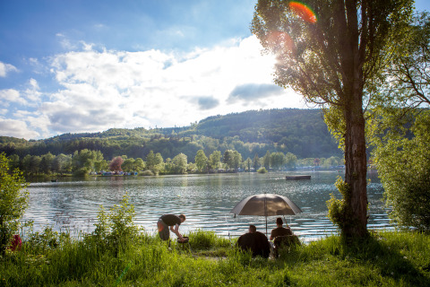 Personas relajándose a la orilla del lago bajo una sombrilla en Camping en Vakantiepark Teichmann, Hessen.