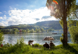 People relaxing by the lakeshore under an umbrella at Camping en Vakantiepark Teichmann, Hessen, Germany.