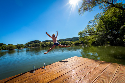 Ragazzo salta dal pontile nel lago al Camping en Vakantiepark Teichmann in una giornata soleggiata, natura intorno.