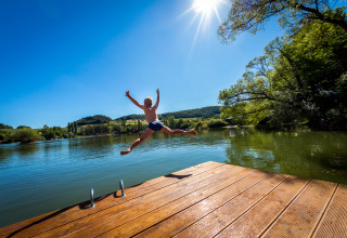 Niño salta desde un muelle al lago en Camping en Vakantiepark Teichmann, día soleado rodeado de naturaleza.