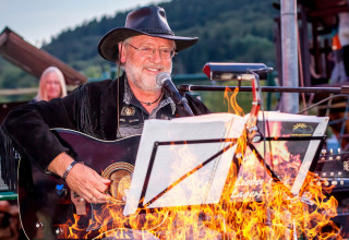 En musiker iført cowboyhat spiller guitar ved et bål under en koncert i Camping en Vakantiepark Teichmann.