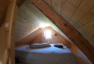 Et hyggeligt soveområde i en træhytte med skrå loft og lille vindue, perfekt til glamping.