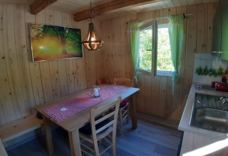 Knusse glamping keuken met houten wanden, eettafel, open raam en groene geruite gordijnen.