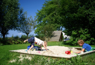 Two children playing in an outdoor sandbox with green trees and It Dreamlân - Ecolodges Friesland in the background.