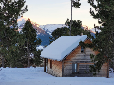 Cabaña de madera con techo nevado en bosque de pinos con vista a las montañas en los Chalets Huttopia, Pirineos.