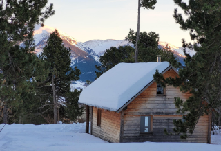 Cabaña de madera con techo nevado en bosque de pinos con vista a las montañas en los Chalets Huttopia, Pirineos.
