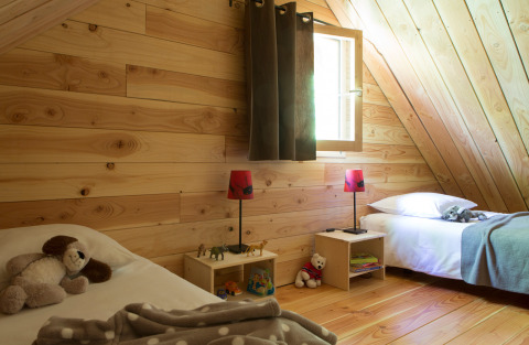 Cozy wooden chalet bedroom with two single beds, toys, lamps, and sunlight streaming through a window.