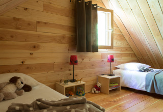Cozy wooden chalet bedroom with two single beds, toys, lamps, and sunlight streaming through a window.