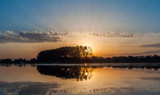 Sonnenuntergang hinter Bäumen an einem See in einem Ferienpark mit Glamping, Wasser spiegelt den Himmel.