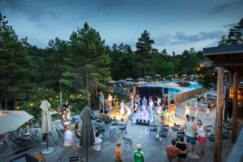 Village Huttopia Lanmary-Périgord : scène de glamping avec invités, estrade, guirlandes et forêt en arrière-plan.