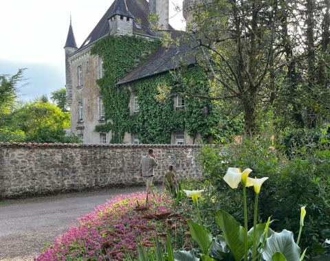 Dos personas caminan junto a un muro de piedra frente a un castillo cubierto de hiedra en Vieflow Lodge Dordogne.