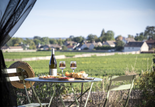Udendørs bord med vin, ost og brød på en glampingplads, med udsigt over vinmarker ved Huttopia Meursault.