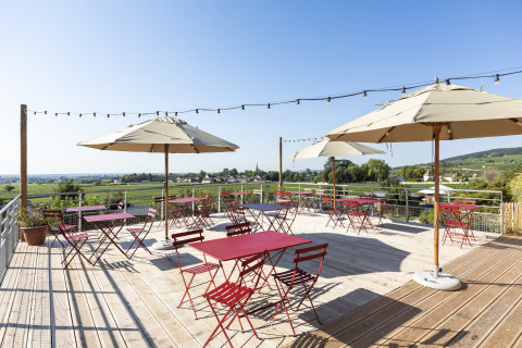 Outdoor terrace at Huttopia Meursault - Glamping Bourgogne with red tables, umbrellas, and a scenic countryside view.