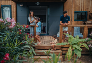 En familie slapper af på terrassen foran deres glamping-hytte omgivet af blomster og grønne planter.