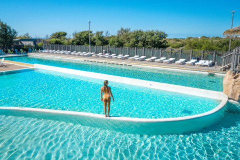 Person in swimwear stands by the pool at Camping Beach Garden - Glamping South France, surrounded by loungers.