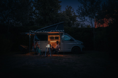 Zwei Menschen entspannen abends vor einem Wohnmobil mit Lichterkette, Camping Beach Garden Glamping.