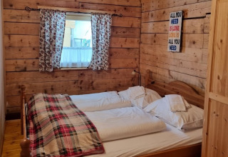 Chambre glamping chaleureuse en bois à Glamping Bad Gastein avec lit double, plaid à carreaux, rideaux fleuris.