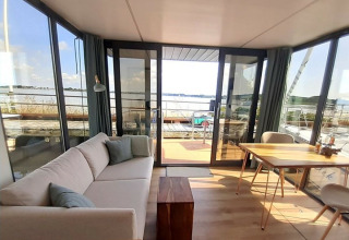 Lichte woonkamer met bank, tafel en panoramisch uitzicht op het meer bij Wind & Wellen - Woontboot Sleeswijk-Holstein.