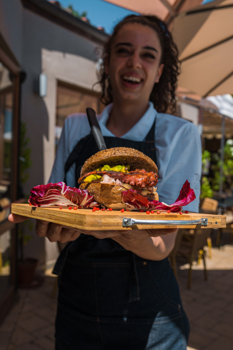Een glimlachende serveerster toont een gourmetburger op een houten plank bij Chianti Glamping Resort buiten.