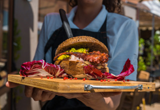 En smilende servitrice fremviser en gourmetburger på en træbakke på Chianti Glamping Resort udendørs.