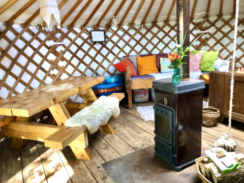Interieur van een yurt met houten tafel, kleurrijke kussens, houtkachel en een gezellige sfeer.