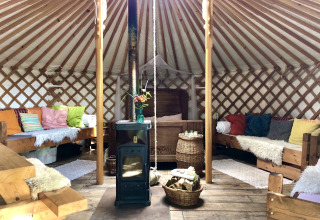 Indretning af en hyggelig yurt med træovn, farverige puder og træmøbler på Glamping 't Buitenland.