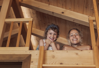 Zwei lächelnde Menschen schauen von einer Holz-Glampinghütte in Camp MondSeeLand, Österreich, nach unten.