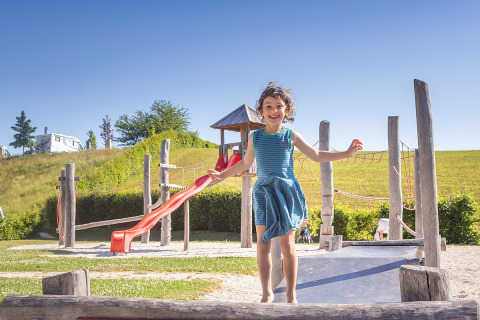 Una niña sonriente juega en un parque con tobogán en Camp MondSeeLand, rodeada de colinas verdes y naturaleza.