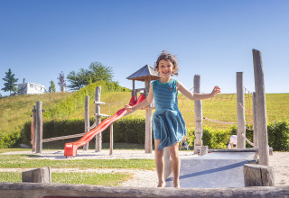 A happy girl plays on a playground with slide at Camp MondSeeLand, surrounded by grassy hills and blue sky.