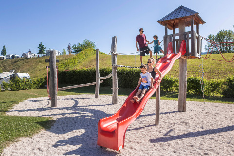 Famiglie si divertono su un parco giochi soleggiato con scivolo al glamping Camp MondSeeLand in Austria.