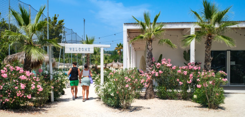 Two people walk into Villa Alwin Beach Resort, surrounded by palm trees and flowers under a blue sky.