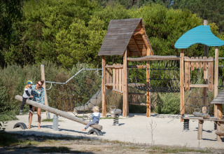 Bambini che giocano su un parco giochi in legno a Huttopia Lagoa de Óbidos glamping, immersi nella natura verde.