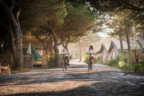 Dos personas pasean en bicicleta por Huttopia Lagoa de Óbidos glamping entre árboles y tiendas en Portugal.