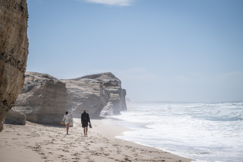 Two people walk on a rocky beach near Huttopia Lagoa de Óbidos - Glamping Portugal, ocean waves nearby.