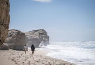 Two people walk on a rocky beach near Huttopia Lagoa de Óbidos - Glamping Portugal, ocean waves nearby.