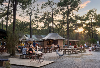 People dining outdoors at Huttopia Lac de Carcans - Glamping Aquitaine, surrounded by pine trees at sunset.