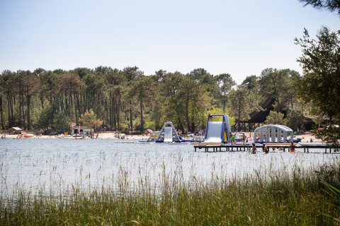 View of a lake with inflatable water structures at Huttopia Lac de Carcans glamping, surrounded by pine forest.