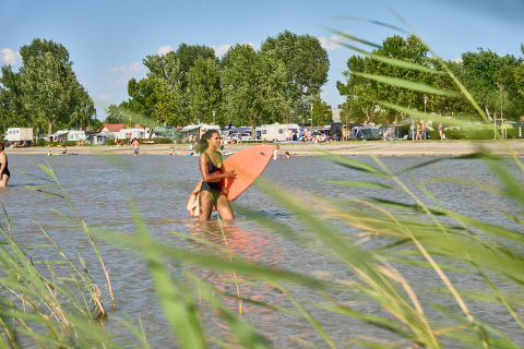 Femme avec une planche de surf au bord du lac au Strandcamping Podersdorf am See, entourée de campeurs et arbres.