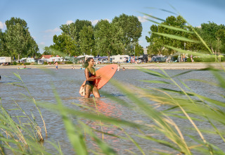 Woman with a surfboard at the lakeshore at Strandcamping Podersdorf am See, surrounded by campers and trees.