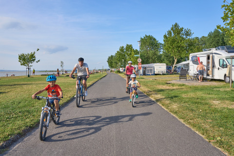 Family cycling on a path at Strandcamping Podersdorf am See - Tiny Houses, with RVs and the lake in the background.