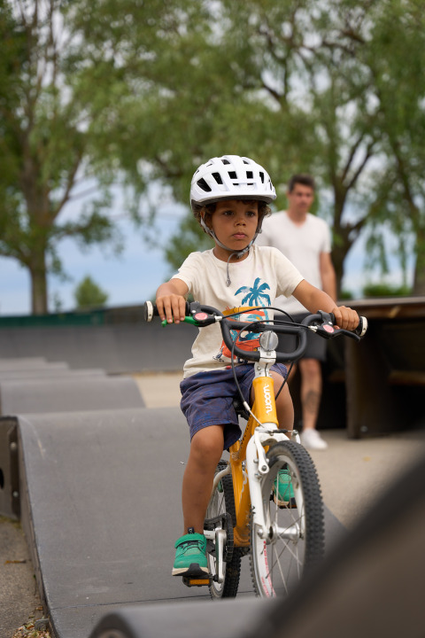 Boy riding bike wearing helmet at Strandcamping Podersdorf am See - Tiny Houses, Austria, adult in background.