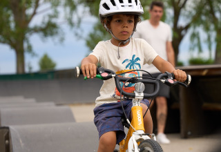 Boy riding bike wearing helmet at Strandcamping Podersdorf am See - Tiny Houses, Austria, adult in background.